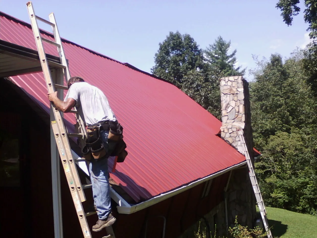 Roofer on a ladder beside a red metal roof and stone chimney — Soffit & Fascia Repair by Roofing Brothers in Saddle River